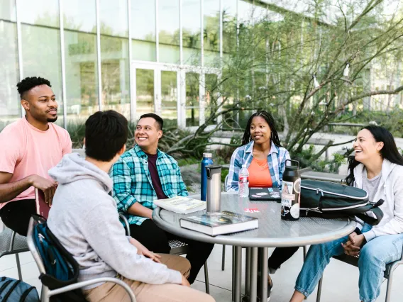 group of students sitting at an outdoor table with books and binders on the table and a building in the background