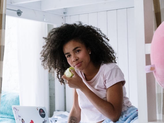 person sitting on a bed eating an apple and looking at a computer