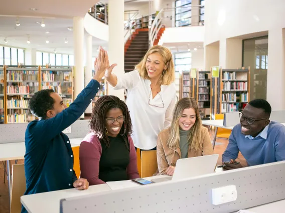 group of students sitting at a desk with one giving a teacher a high five