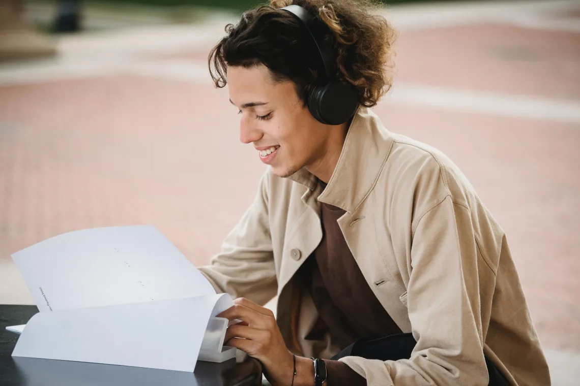 person sitting alone at a table wearing headphones and reading from a stack of papers