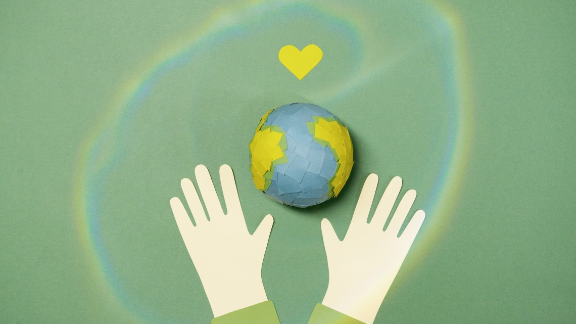 paper cutout of two hands open around a paper earth with a paper heart above it
