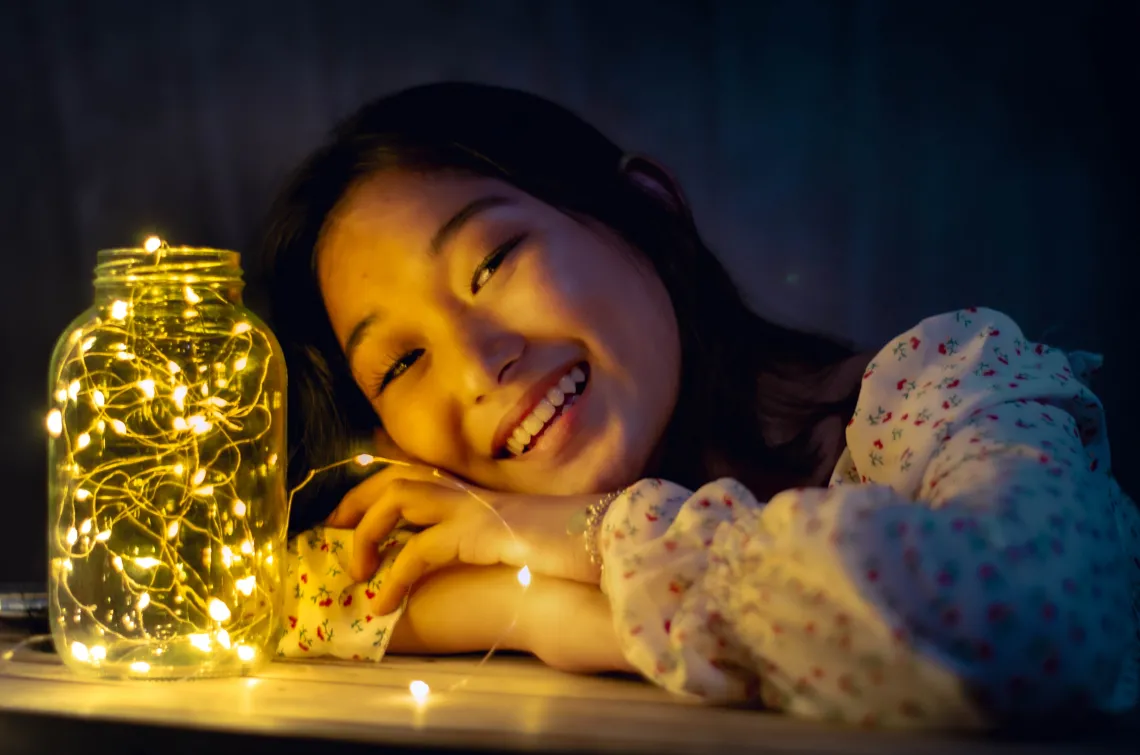 person resting their head on a desk with a jar filled with twinkle lights nearby