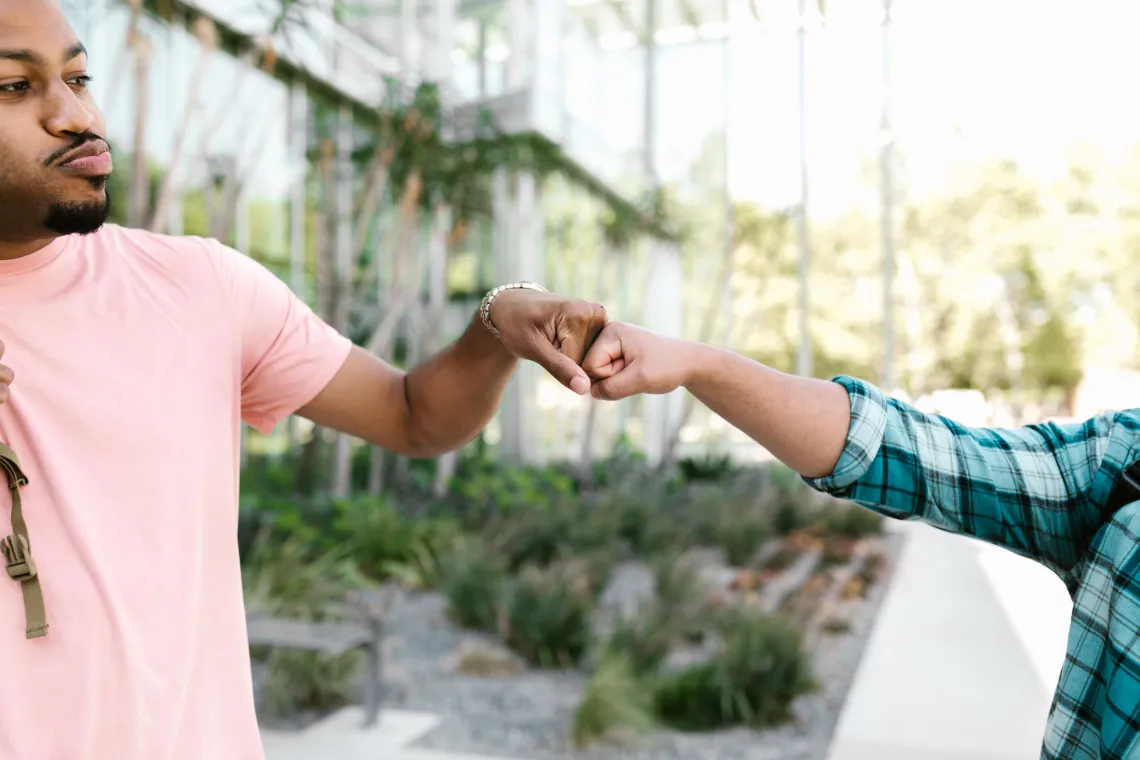 two people bumping fists outdoors with a glass building and trees in the background